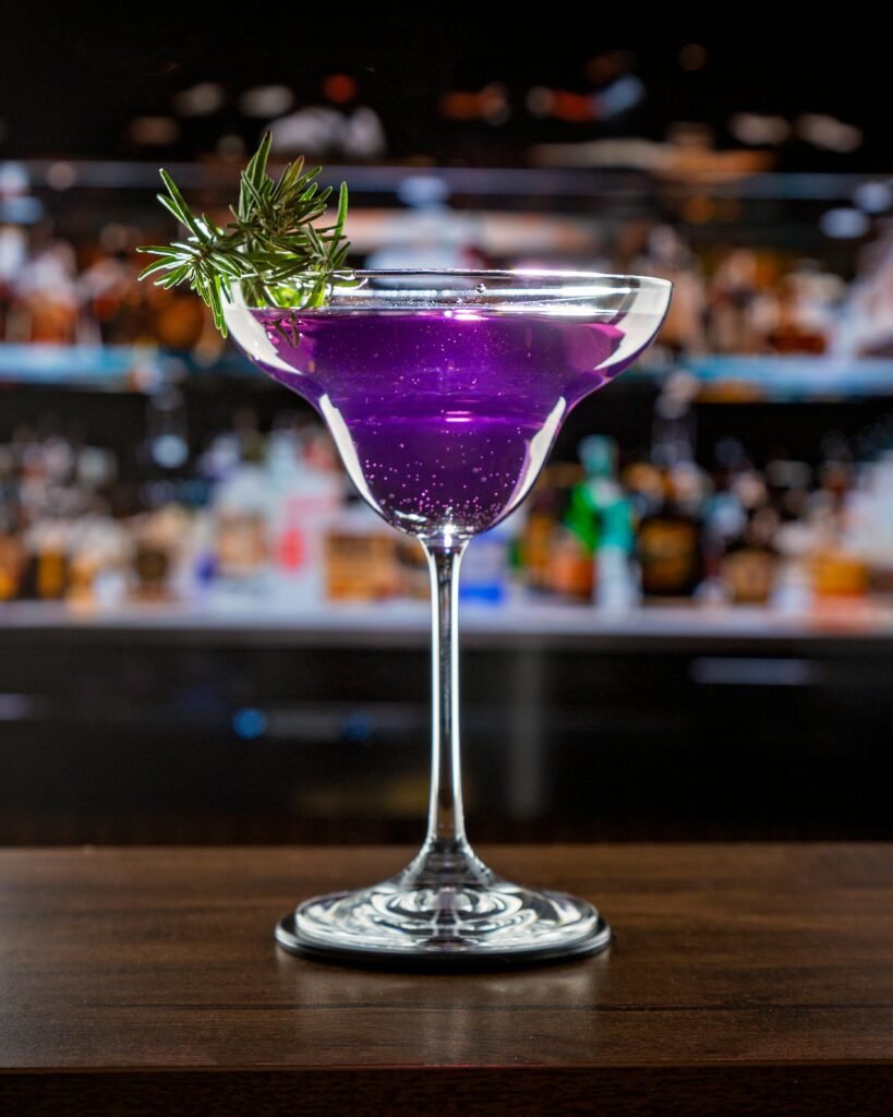 Vibrant purple cocktail with rosemary garnish on bar counter, perfect for nightlife scenes.